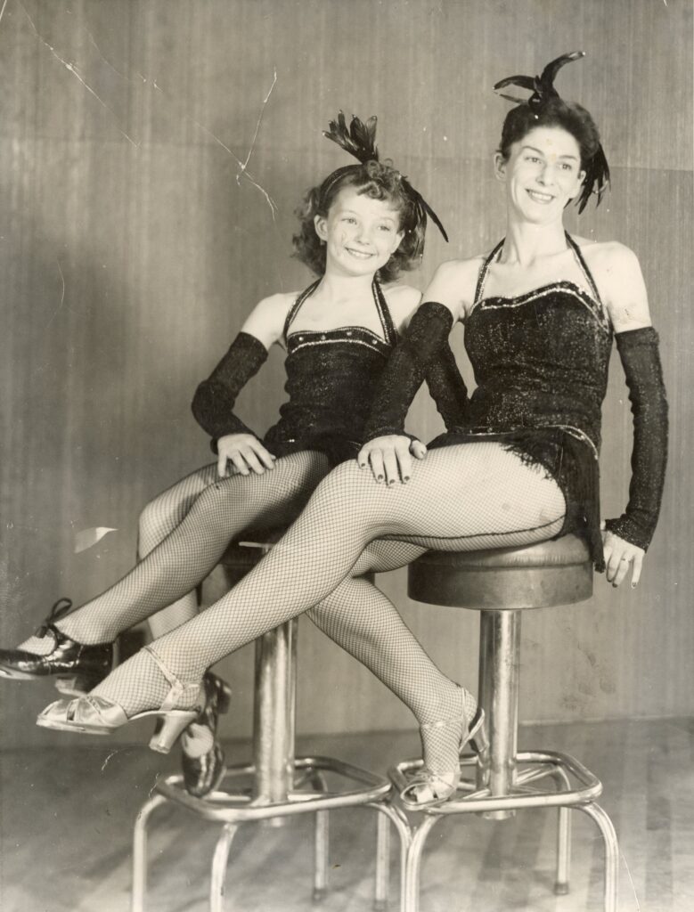 Polly Nulty and her daughter Mary in matching dance costumes and tap shoes pose on stools.