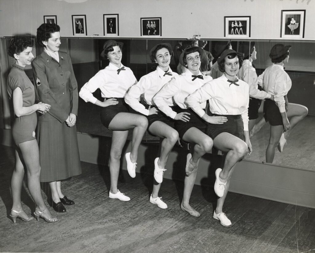 A dance instructor and a woman drill seargeant in uniform look at four you women dancers practicing in the Nulty studio.
