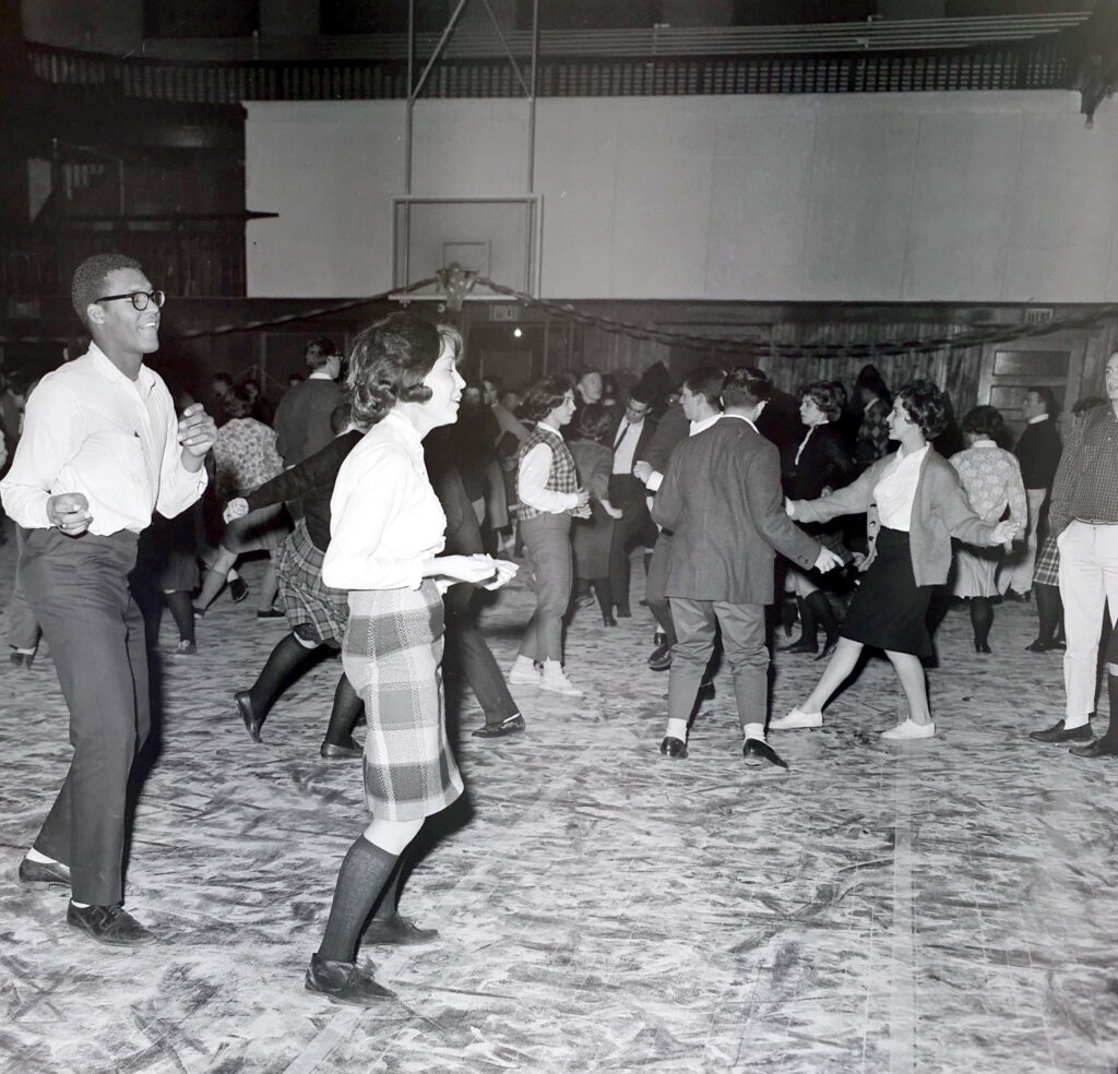 Photograph of a male and female student walking in the gym while smiling and snapping their fingers. Other students dance in the background.