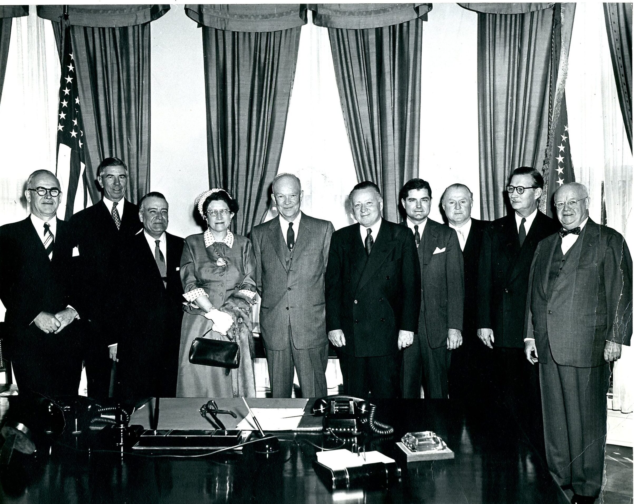 A group portrait showing several men wearing suits as well as Consuelo Northrop Bailey and President Eisenhower in the center. Everyone is looking at the camera and smiling. The President’s desk is visible in the foreground while two flags and several window treatments are apparent in the background.