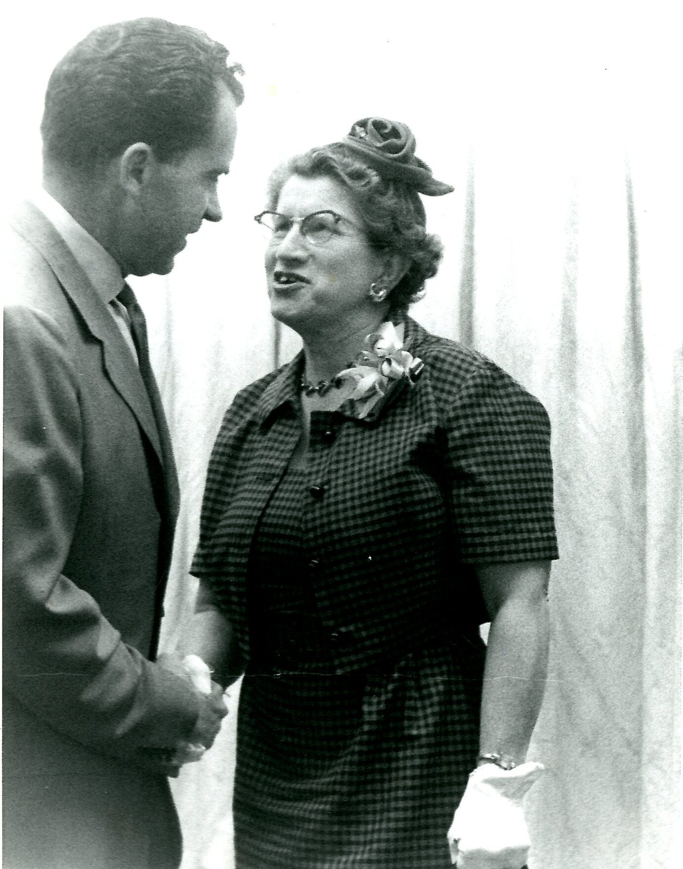 A photograph of Richard Nixon, visible in an obscured profile, stood shaking hands with Consuelo Northrop Bailey in front of a light-colored curtain. She appears to be speaking and he is gently smiling; they are both making eye contact with each other.
