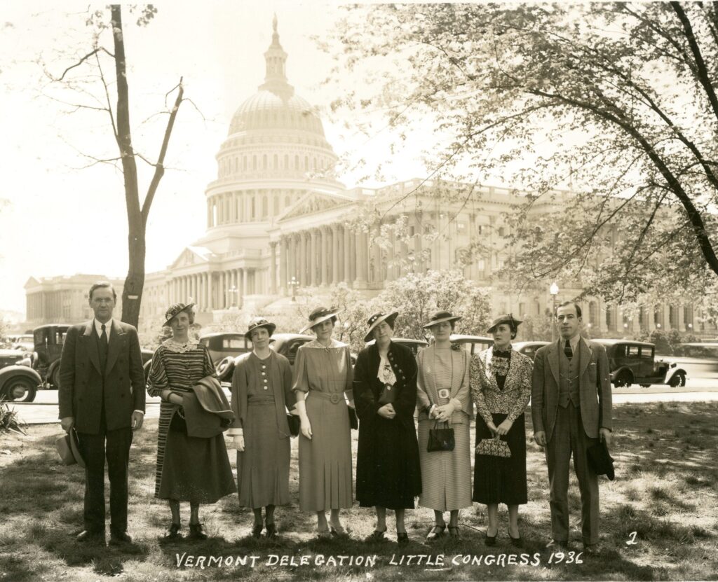 A photograph of eight adults (six women flanked by two men) standing shoulder to shoulder with the capitol building in Washington, D.C. in the background The men wear dark three piece suits with ties and hold their hats while the women wear skirts suits or long sleeved dresses, hats, and gloves.