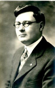 A formal portrait of a man wearing a suit and round glasses