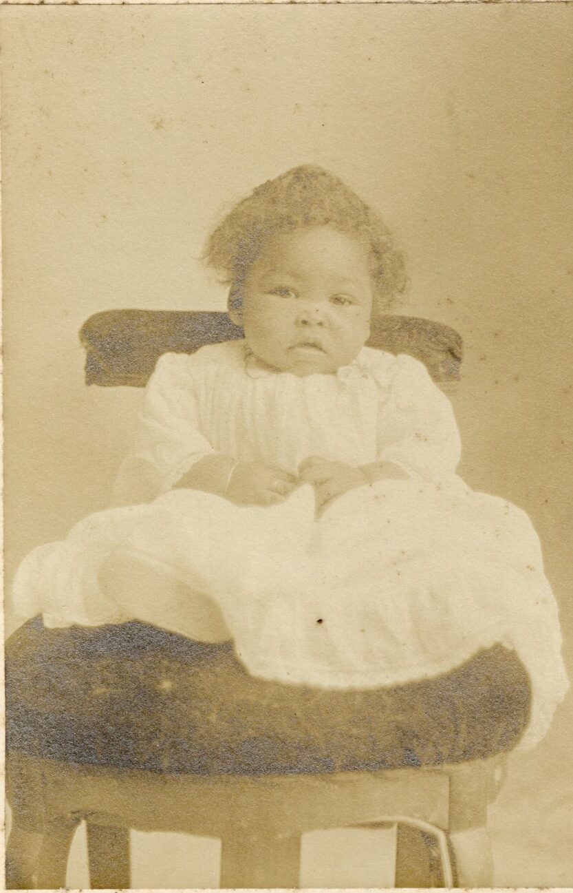Studio portrait of a young black child seated on a chair and possibly wearing a white christening outfit.