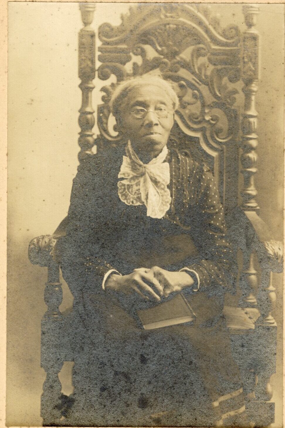 A studio photograph shows an older Black woman sitting in an ornate chair. her hands are folded over a book in her lap.