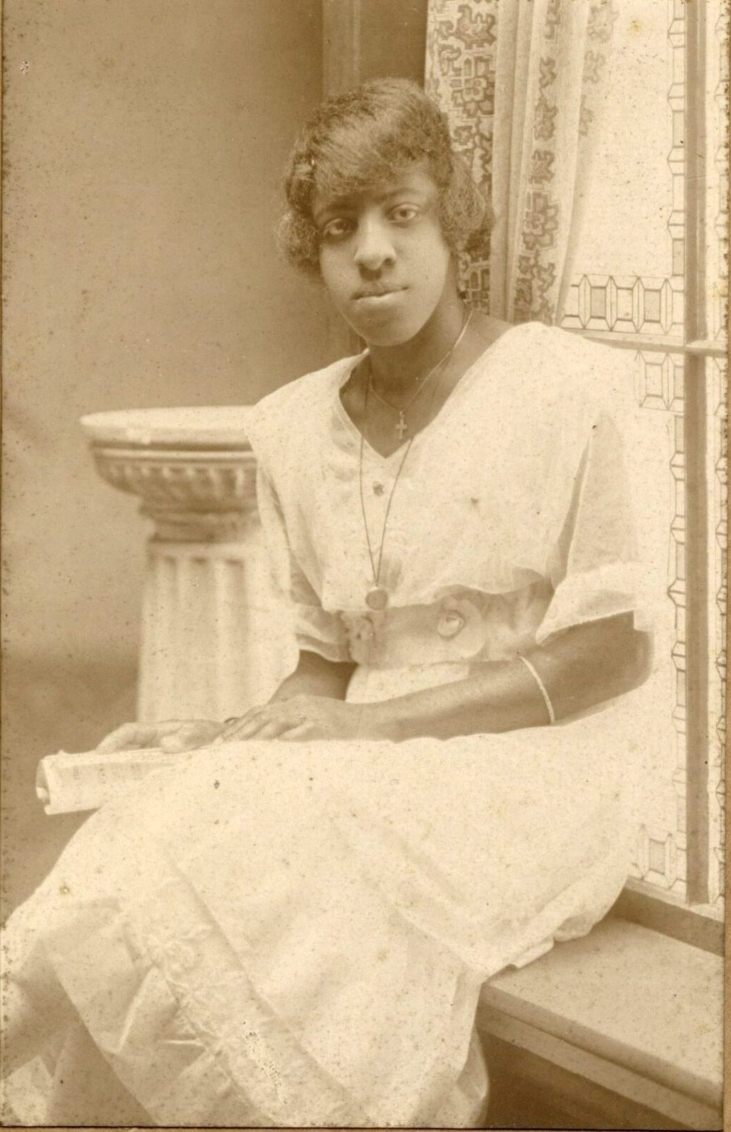 Studio photograph of a young Black woman sitting on a ledge in front of window. She is wearing a fancy white dress and may be holding a diploma.