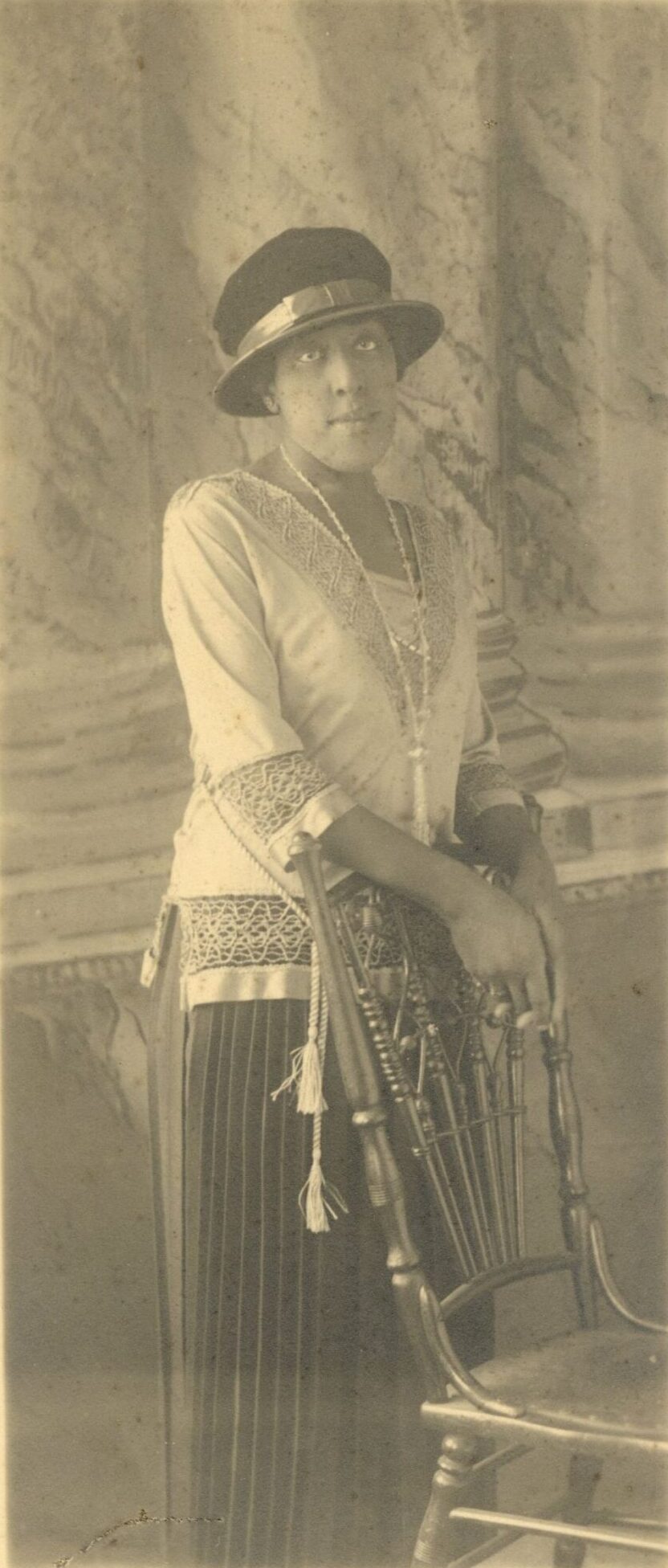 This studio photograph shows a young Black woman standing next to a chair. She is wearing a hat, an embroidered top and a long pleated skirt. 