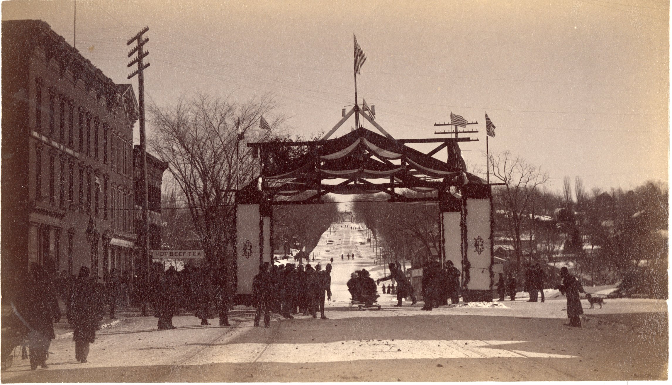 Phot shows the Main Street coasting hill, a traverse sled about to cross under the Coasting Club arch where spectators are gathered..