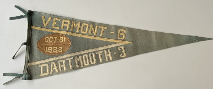 Photo shows a faded green triangular pennant with felt letters "Vermont-6" on the top and "Dartmouth-3" appliqued on the bottom of a felt V. A leather football appliqued in the wide part of the V bears the date Oct. 21 1922 .