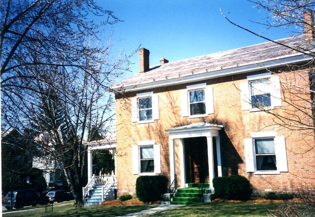 Photo of a brick house with white shutters, end chimneys, a front porch and a side porch.
