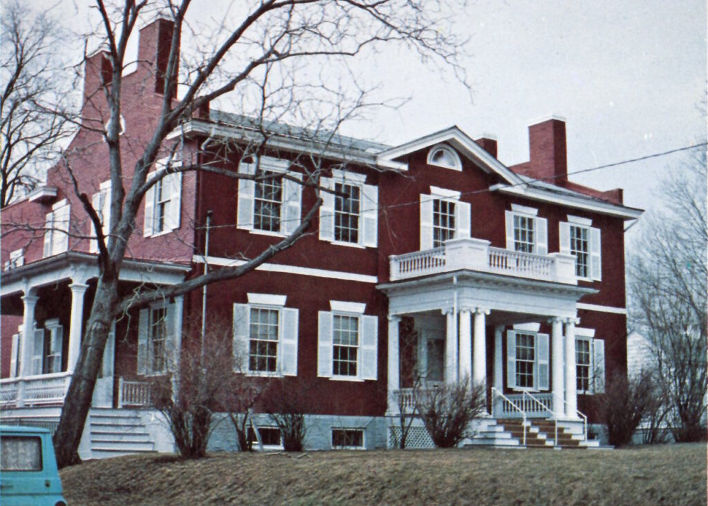 Photograph of large brick house with white shutters, two porches with columns, and end chimneys.