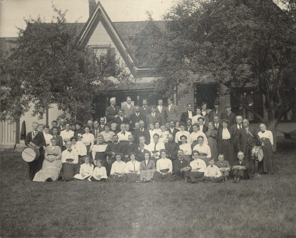 Large group of people of all ages seated and standing in the yard in front of a frame house.