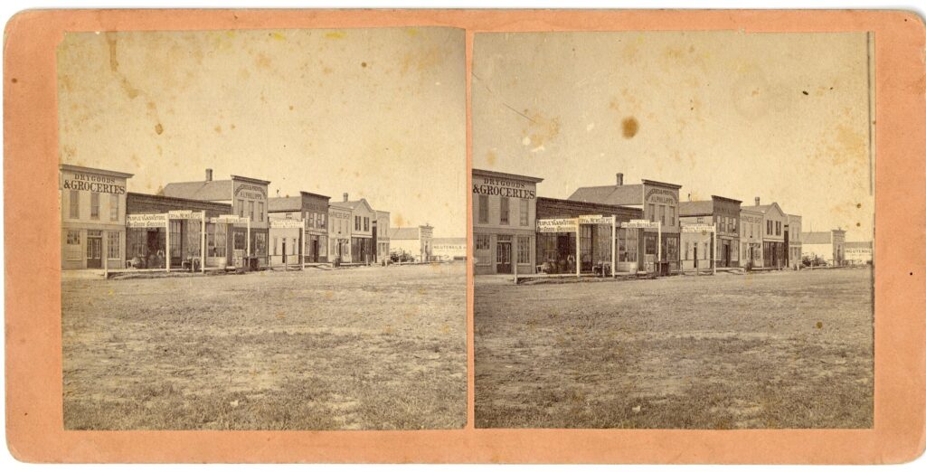 Two photographs mounted side by side on a pink card showing commercial buildings facing a dirt street.