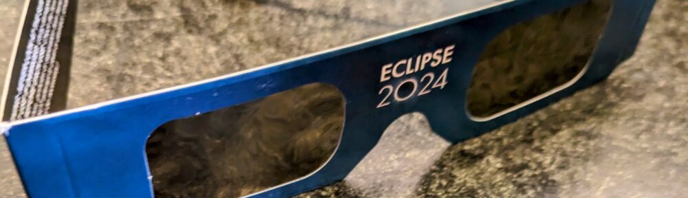 A pair of paper cardboard eclipse glasses in blue with Eclipse 2024 noted