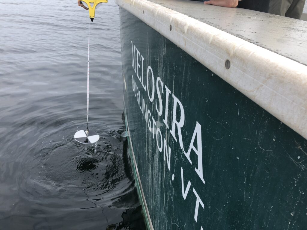 Photo of someone lowering a Secchi disc to assess water clarity off the stern of the R/V Melosira, the University of Vermont's research vessel. 
