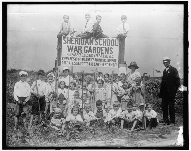 children pose around a sign that reads "Sheridan School War Gardens.".There is an adult standing on the edge of the image.