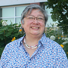 Photograph of Cathy, she is smiling, wearing glasses and a blue and white floral shirt, she has short grey hair.