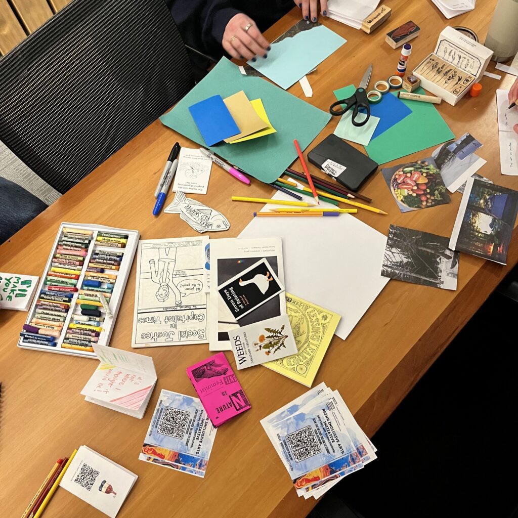 Photo taken from above of a table covered in art supplies at a Zine workshop. The supplies includes paper, glue, pencils, pastels, pens, collage materials, and completed zines.