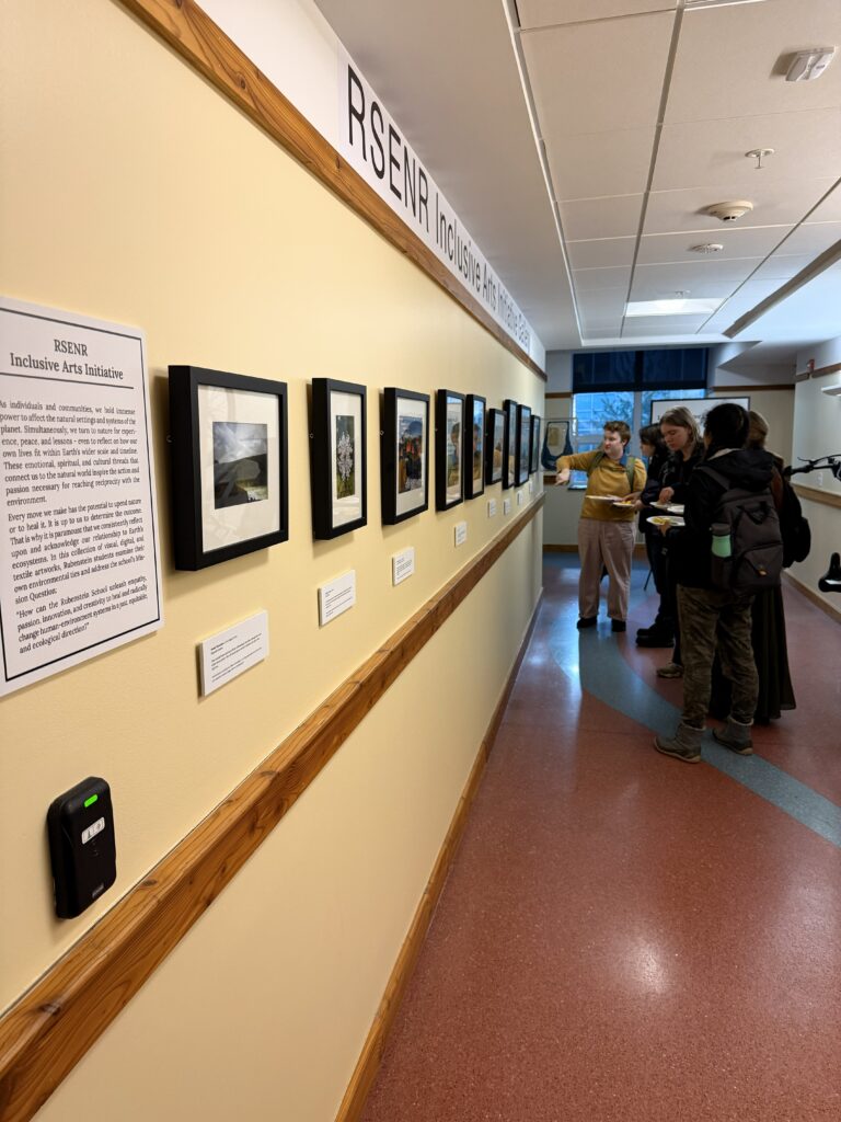 Photograph of people standing in a hallway looking at framed pieces of artwork on a wall. The people are holding paper plates with food on them. The event is a gallery opening in the Aiken Center at UVM.