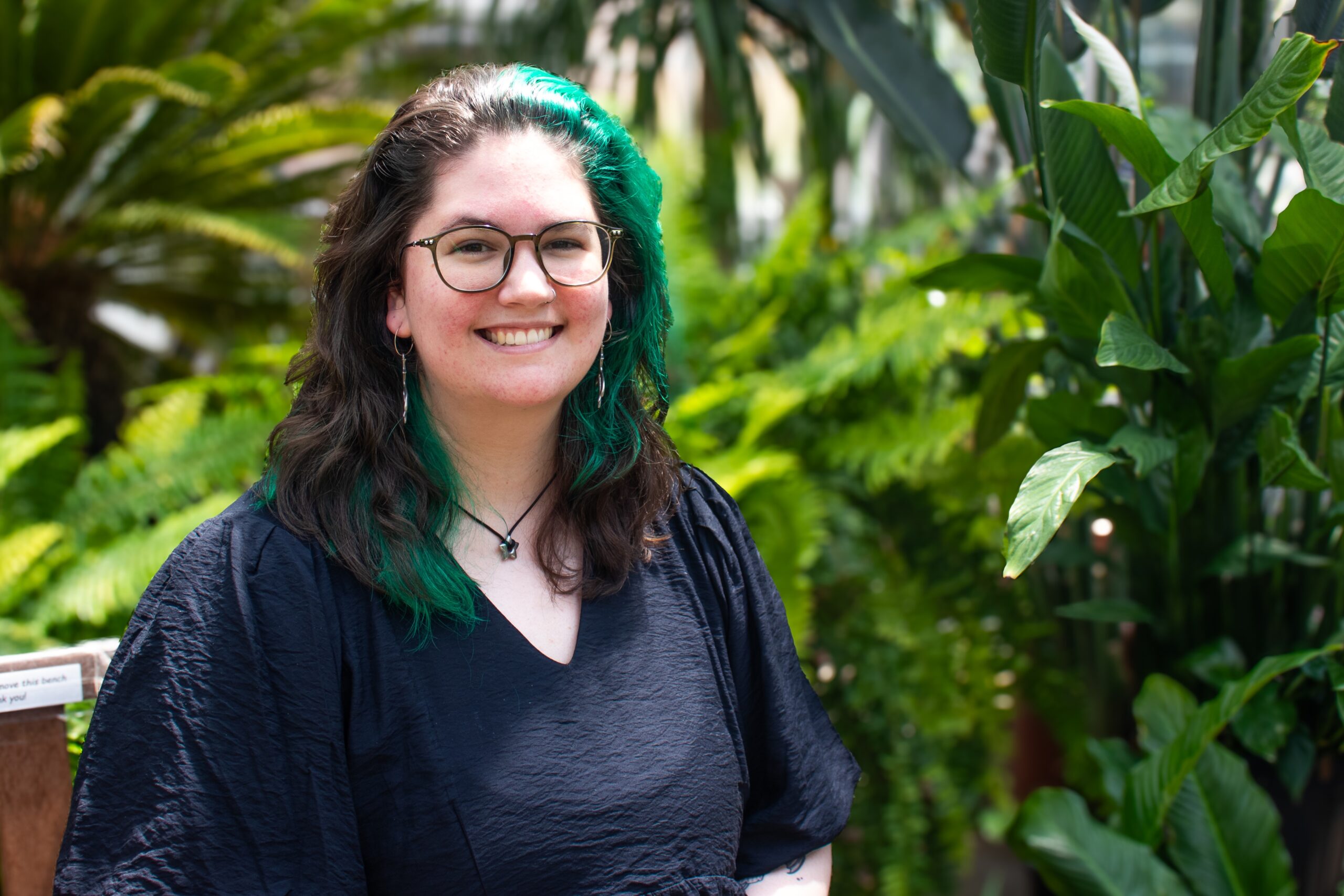 Photograph of Hannah, they are smiling, wearing glasses and a black shirt, they have brown and green hair.