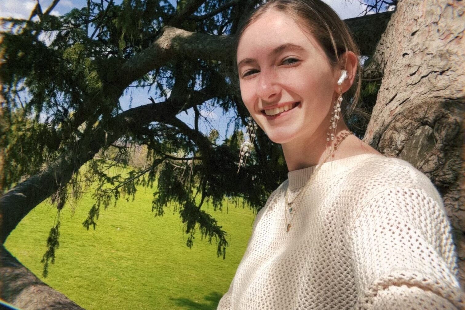 Smiling young woman in a cream sweater sits beneath a sprawling tree, holding a tablet, with a sunny green park and blue sky behind her.