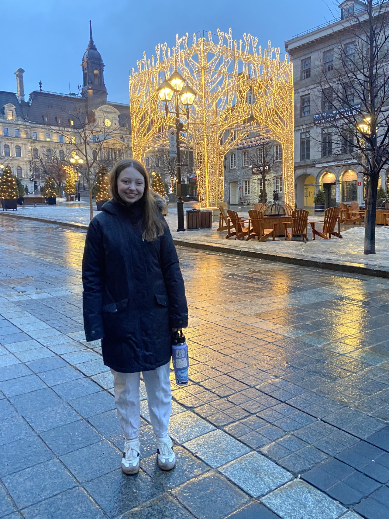 Smiling woman in a dark coat stands in a wet, snowy plaza beneath glowing holiday lights, with historic buildings and outdoor cafés behind her.